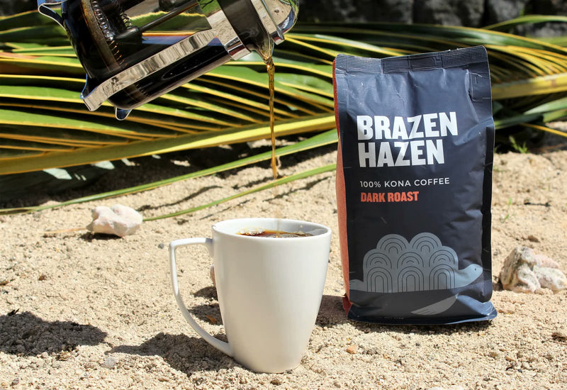 Fresh Brazen Hazen dark roast coffee pouring from a French press into a white mug beside the bag